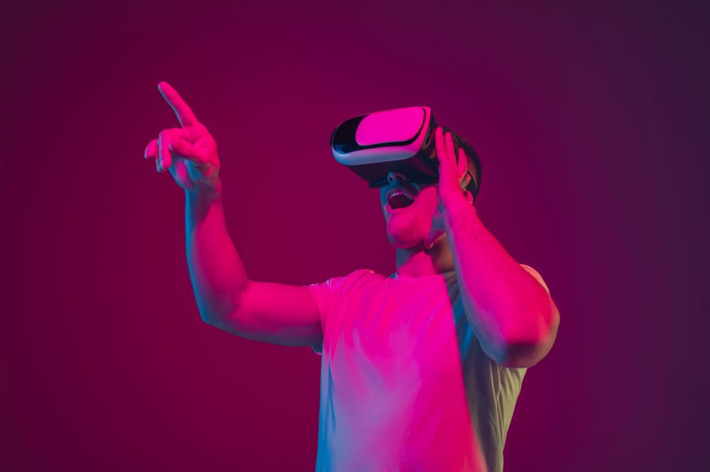 Playing with VR, shoting, driving. Caucasian man's portrait isolated on pink-purple studio background in neon light. Male model with devices. Concept of human emotions, facial expression, sales, ad.